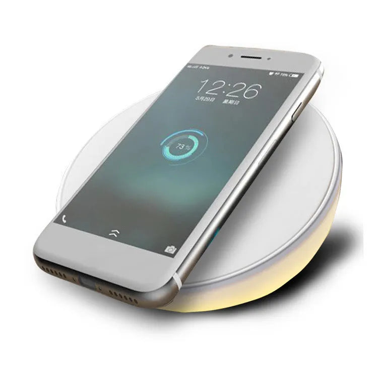 White Qi wireless charger with digital clock display on a desk next to a mobile phone.