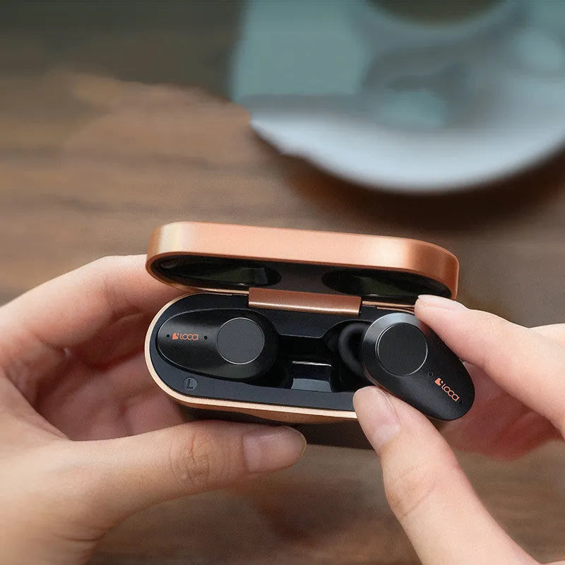 Black wireless Bluetooth headset with bilateral earpieces and a charging stand on a table.