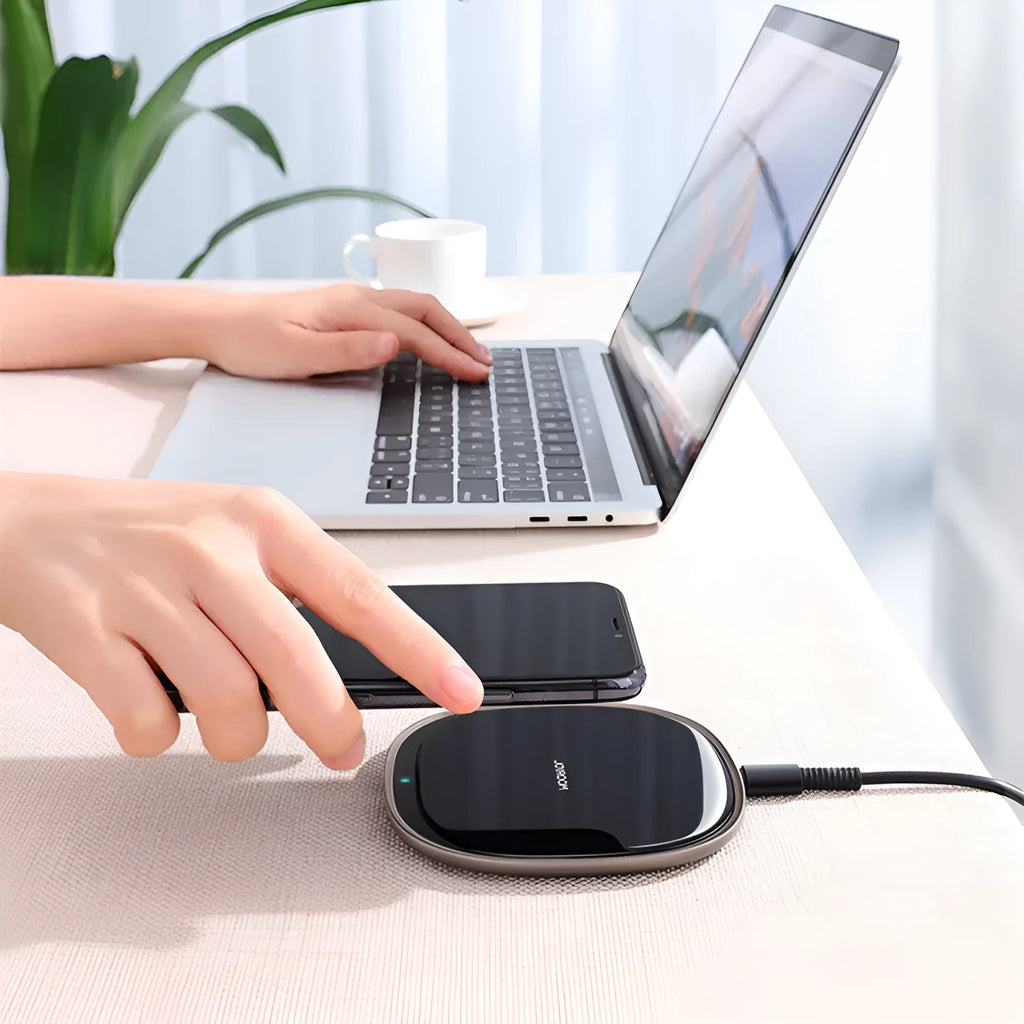 Black 15W wireless phone charger on a wooden desk next to a mobile phone.