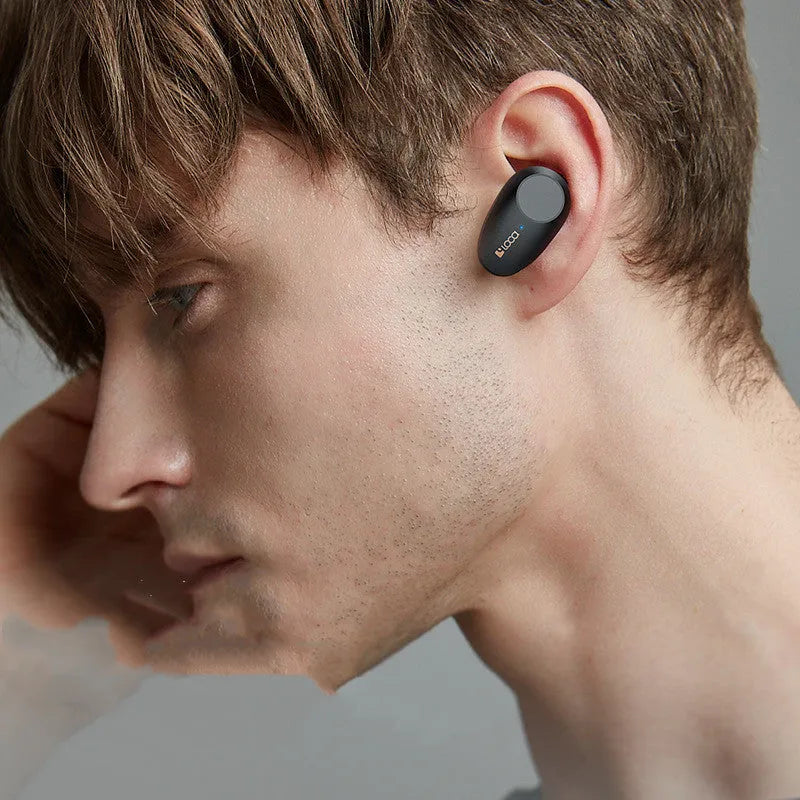 Black wireless Bluetooth headset with bilateral earpieces and a charging stand on a table.