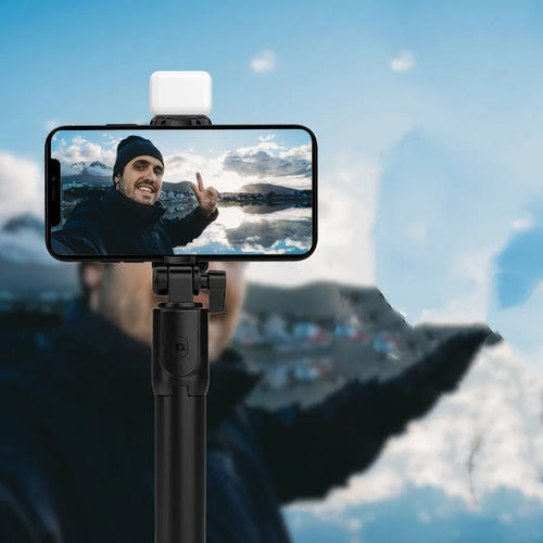 Black Bluetooth selfie stick tripod with phone holder, fill light, and remote control on a table.
