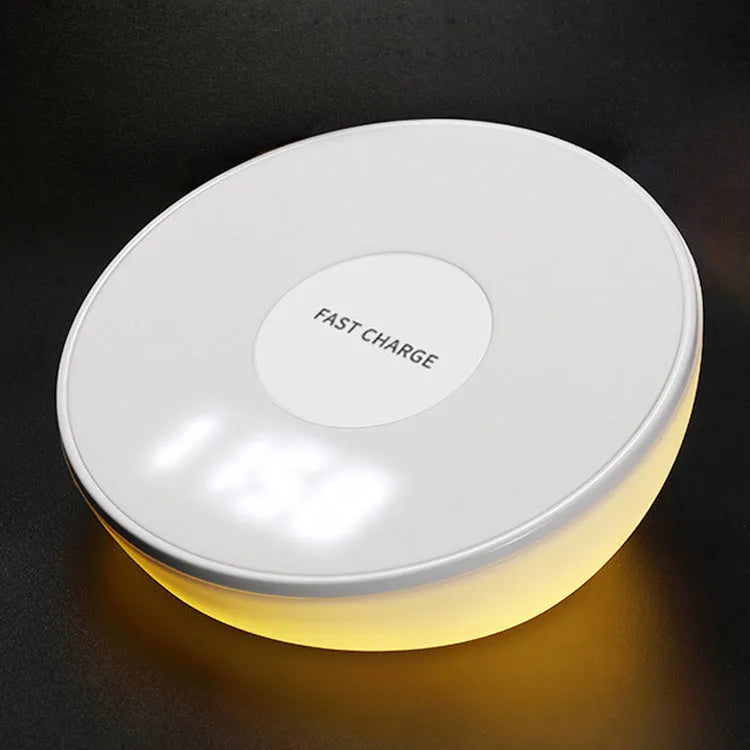 White Qi wireless charger with digital clock display on a desk next to a mobile phone.