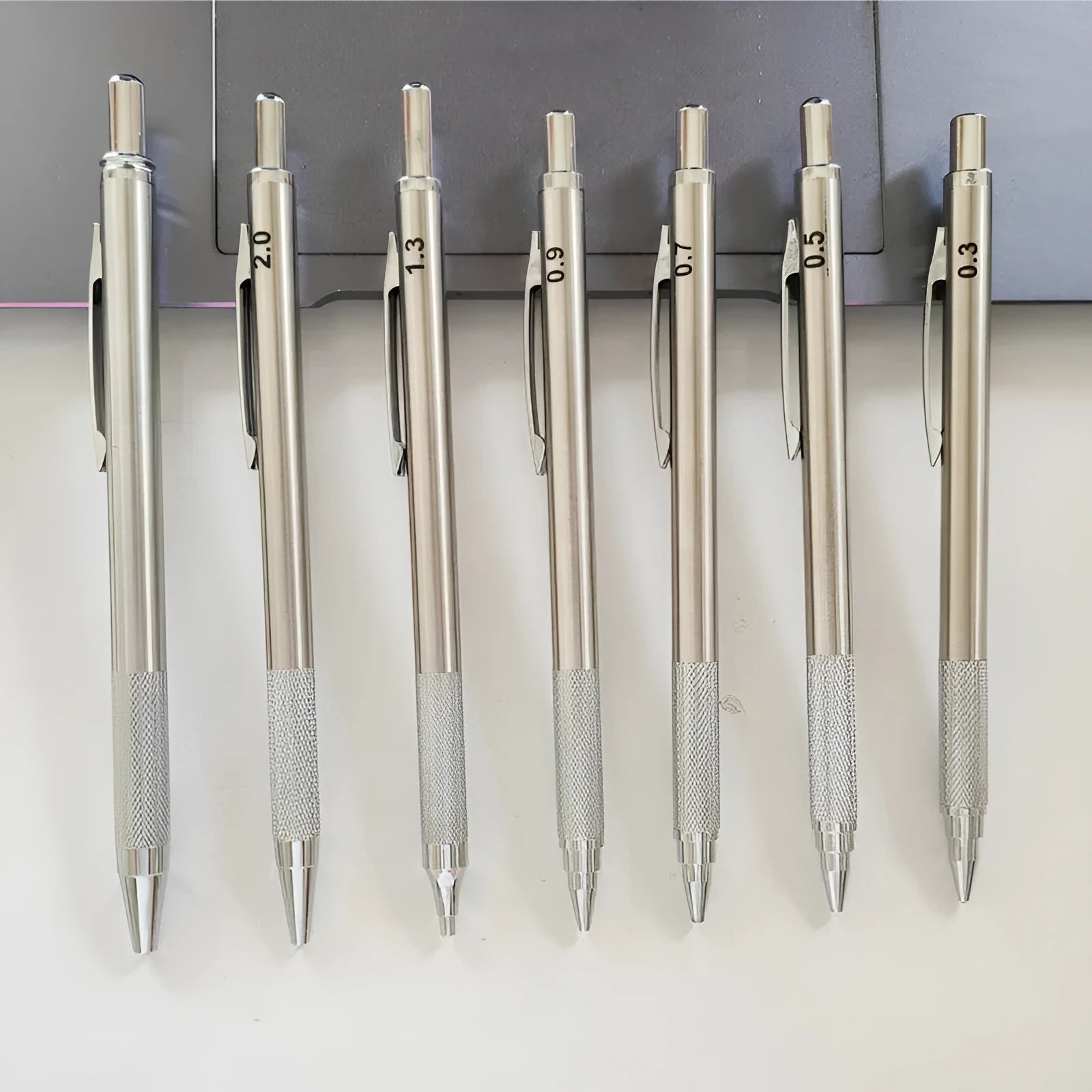Metal automatic mechanical pencil on a surface, shown with multiple lead size indicators including 0.5mm and 2.0mm.