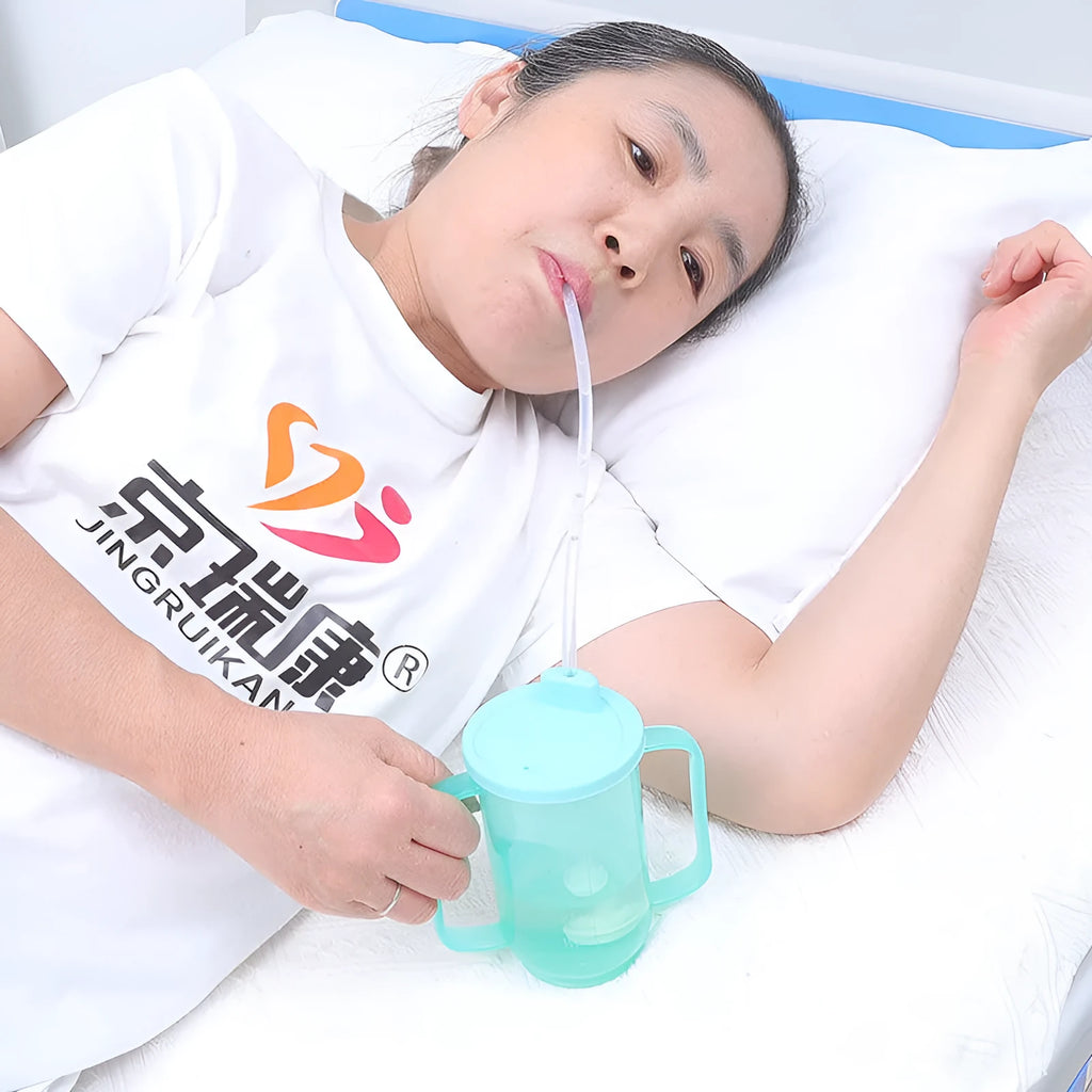 Adult bedside drinking cup with angled choke-proof mouth, handle, and lid, shown in care setting.