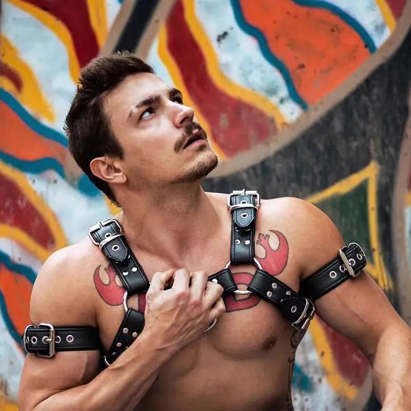 Black leather chest harness strap with a metal buckle and rivets on a neutral background.