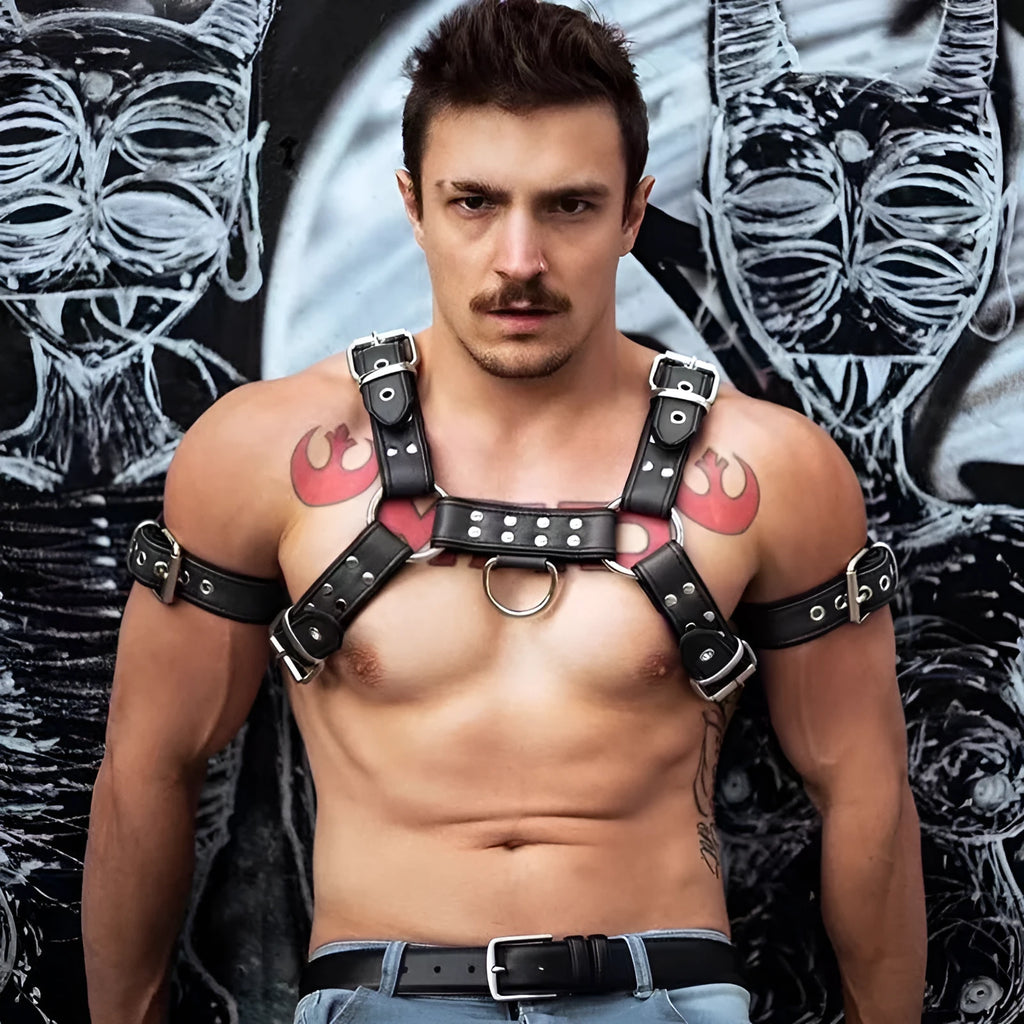 Black leather chest harness strap with a metal buckle and rivets on a neutral background.