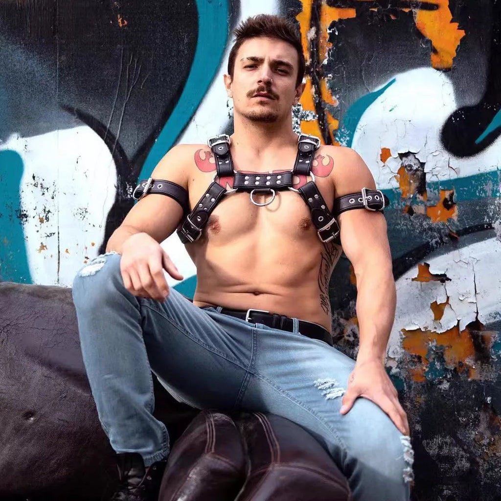 Black leather chest harness strap with a metal buckle and rivets on a neutral background.