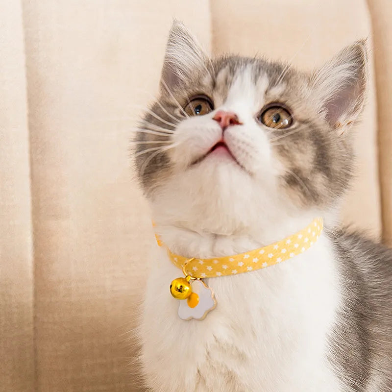 Green adjustable cat collar with a metal bell and a pink bow tie attached, shown on a measuring tape indicating size