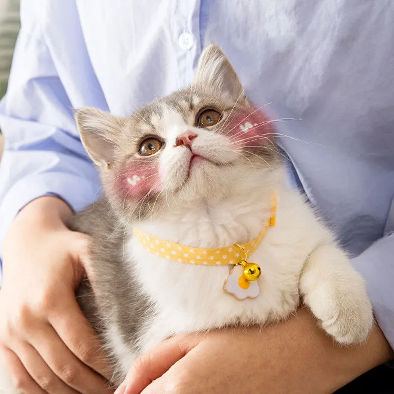 Green adjustable cat collar with a metal bell and a pink bow tie attached, shown on a measuring tape indicating size