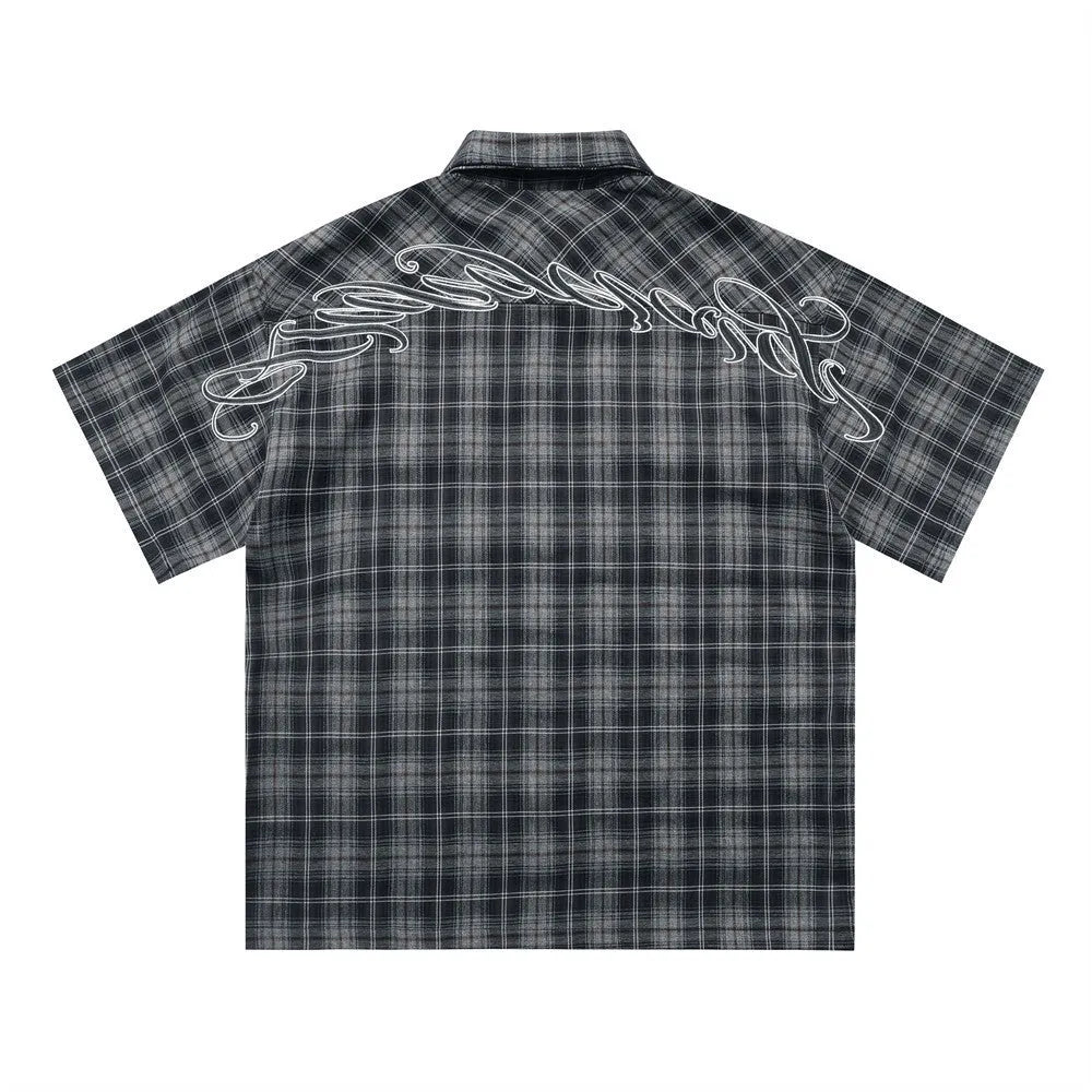 Man wearing a black and brown plaid short sleeve shirt with embroidered letters on the lapel