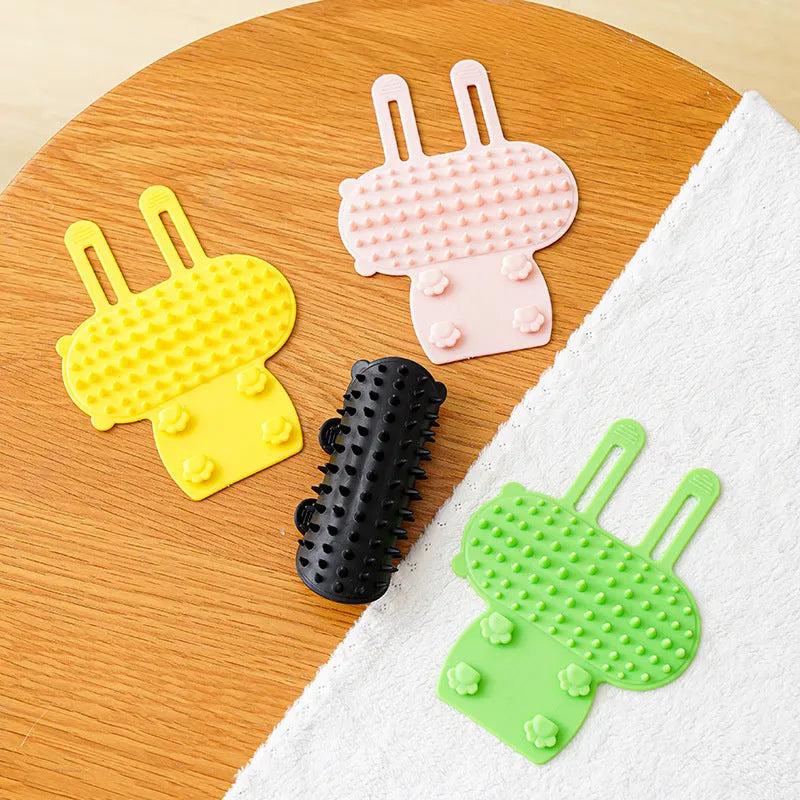 Silicone cat grooming comb with adjustable strap mounted on a table leg, shown in assorted colours.